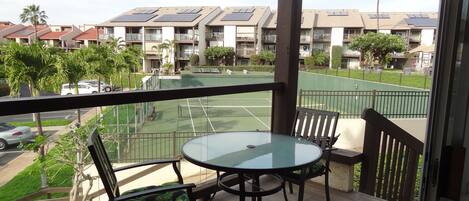 Un balcón con una mesa y sillas de vidrio, con vista a una cancha de tenis y una fila de edificios con paneles solares.
