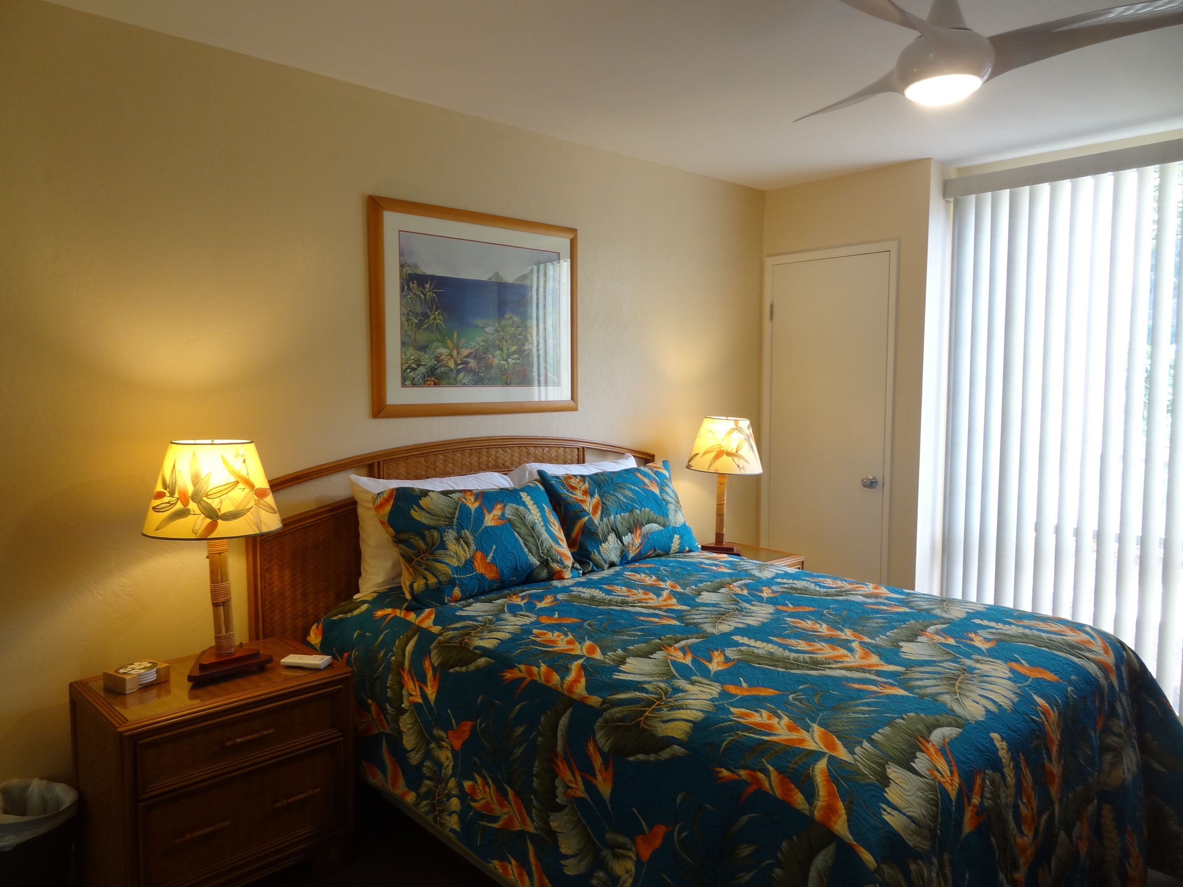 Una cama bien hecha con ropa de cama floral, dos lámparas de noche, una mesita de noche de madera, un cuadro en la pared y una ventana con persianas.