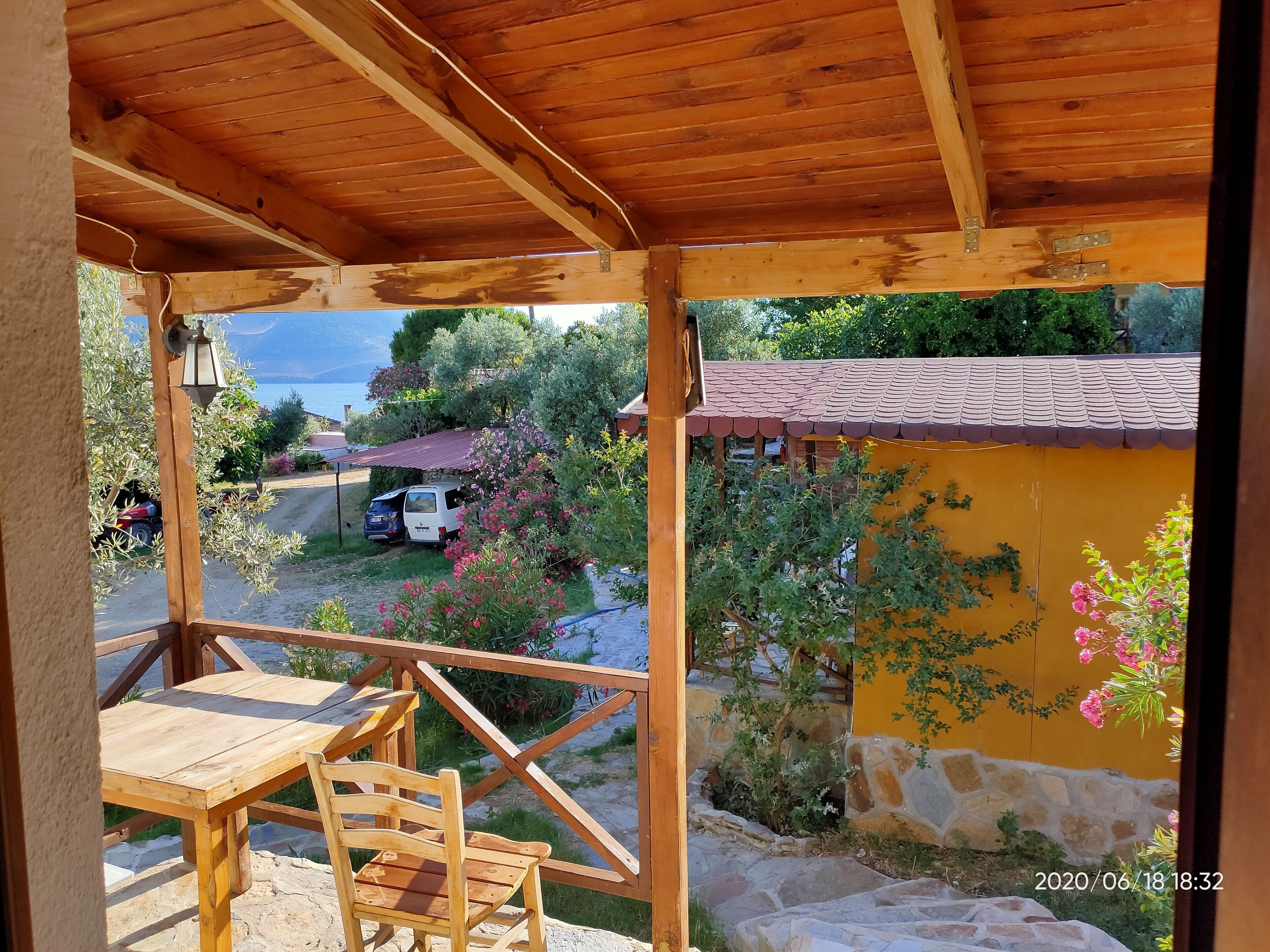 A wooden pergola with a table and chair, overlooking a garden with a yellow wall and pink flowers.