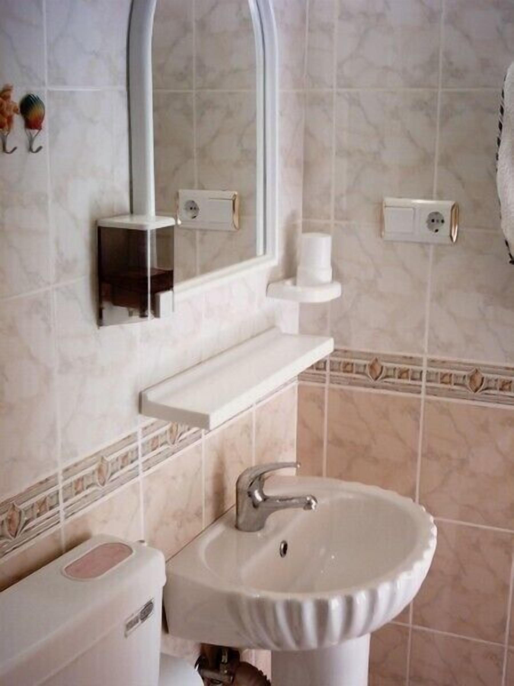 Un baño con lavamanos blanco, un espejo y un inodoro. Hay dos tomas de corriente en la pared.