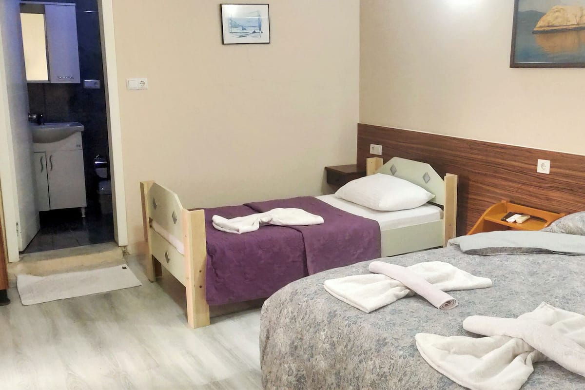 Quarto de hotel com uma cama, uma pequena mesa de trabalho, uma cadeira e um banheiro visível através de uma porta aberta.