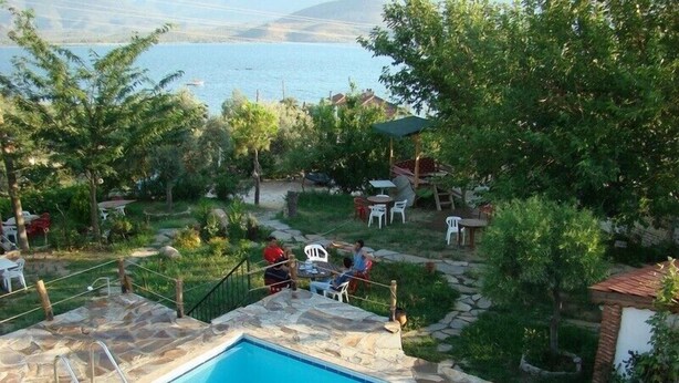 A swimming pool with a stone border, surrounded by a garden with trees and outdoor furniture. - Selenes Pansiyon (Milas)