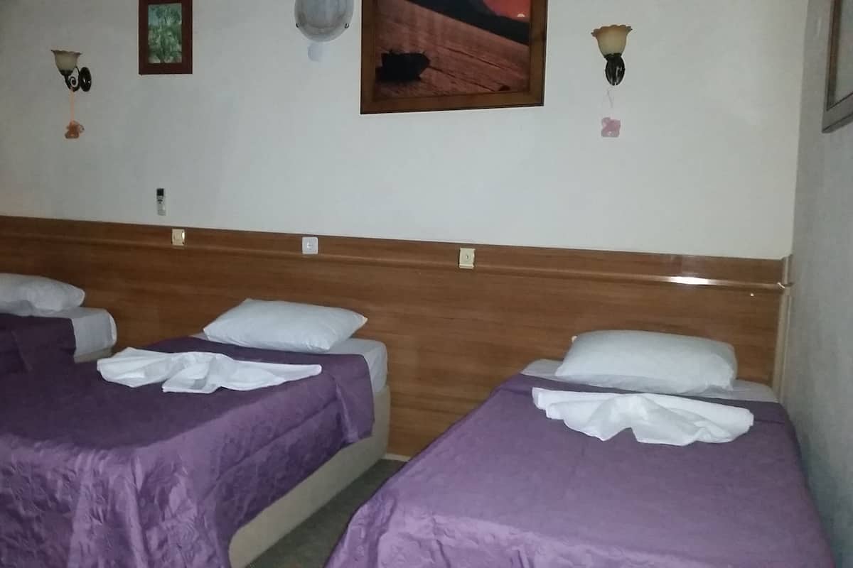 Quarto com três camas, cada uma com colcha roxa e almofadas brancas. Há luminárias fixadas na parede e um quadro emoldurado.