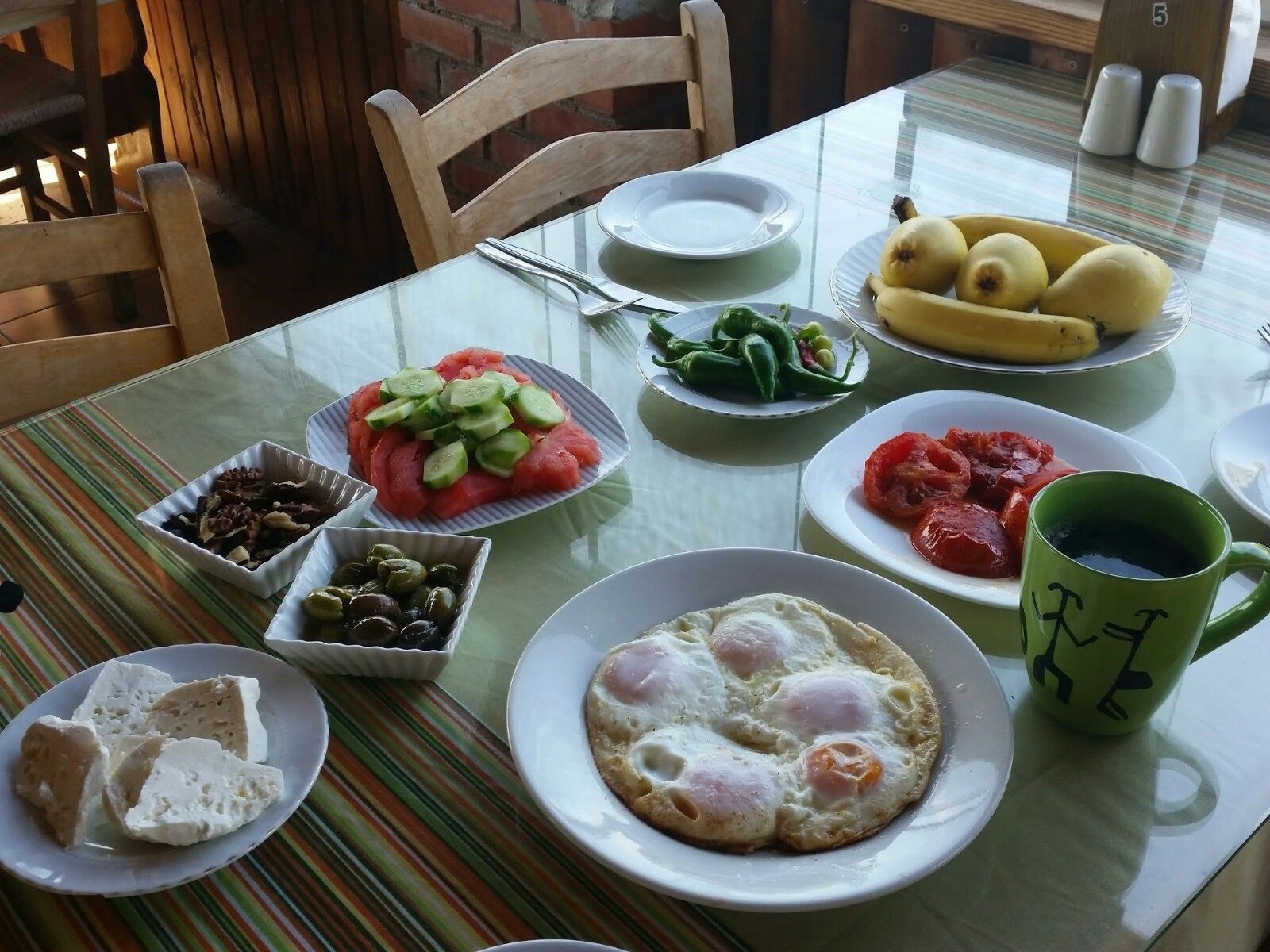 Una mesa de desayuno con platos de comida, entre ellos huevos, tomates, pepinos y una banana.