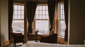 Habitación de hotel con un ventanal grande, una cama, dos sillas y una mesita con un tazón y vasos.