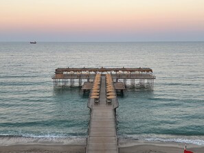 Un muelle que lleva a una cabaña sobre el agua, con sombrillas de paja y mesas de madera.