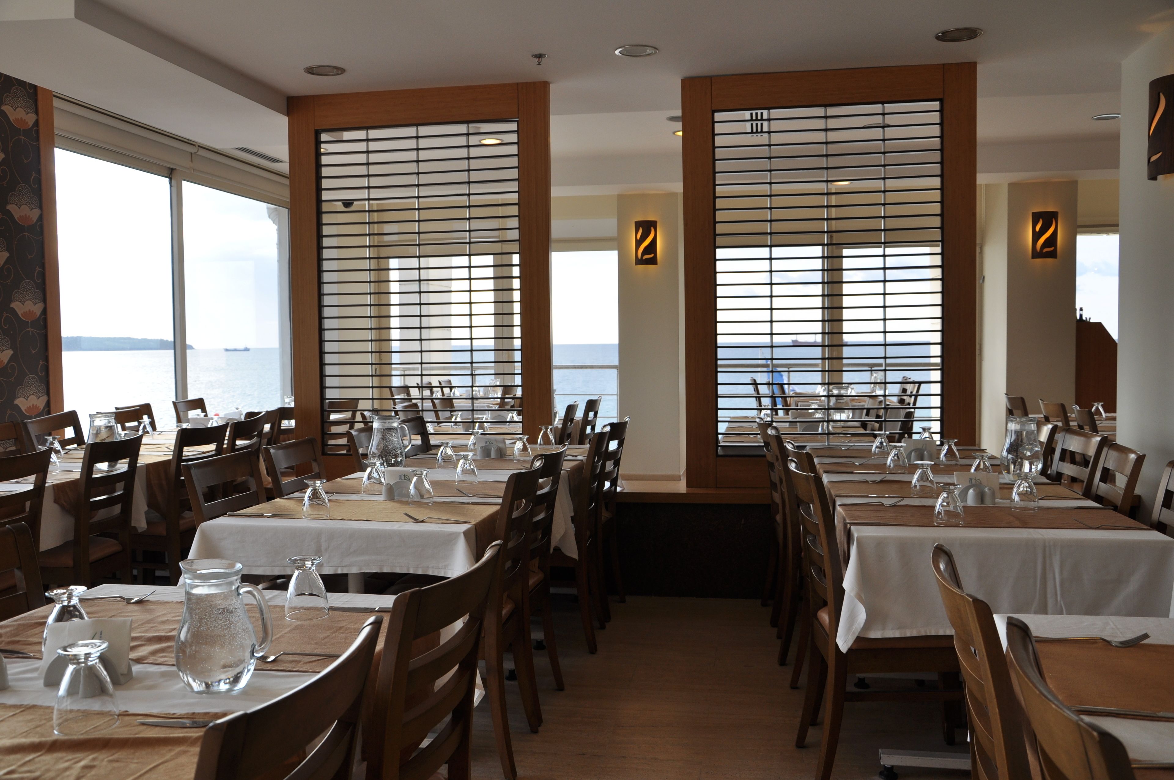 Un restaurant avec de nombreuses tables dressées pour le service, de grandes fenêtres équipées de stores et une vue sur la mer.