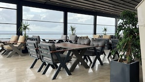 A modern outdoor seating area with tables and chairs, large windows offering a view of the sea, and potted plants. - Igneada Resort Hotel & Spa  (Demirköy)