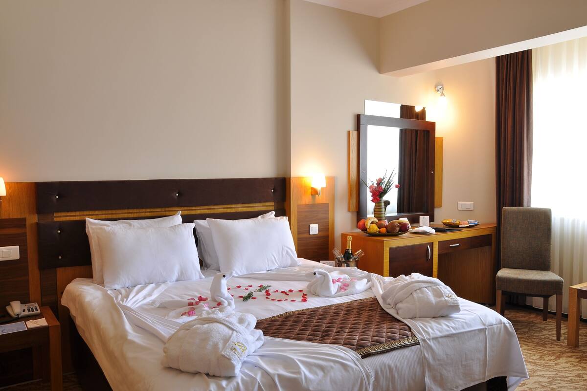 Quarto de hotel com uma cama grande, cabeceira de madeira, escrivaninha com cadeira, espelho e mesa de cabeceira com telefone.