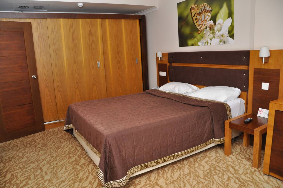 Quarto de hotel com um guarda-roupa de madeira grande, uma cama com colcha marrom, uma mesa de cabeceira com um controle remoto e uma luminária fixada na parede.