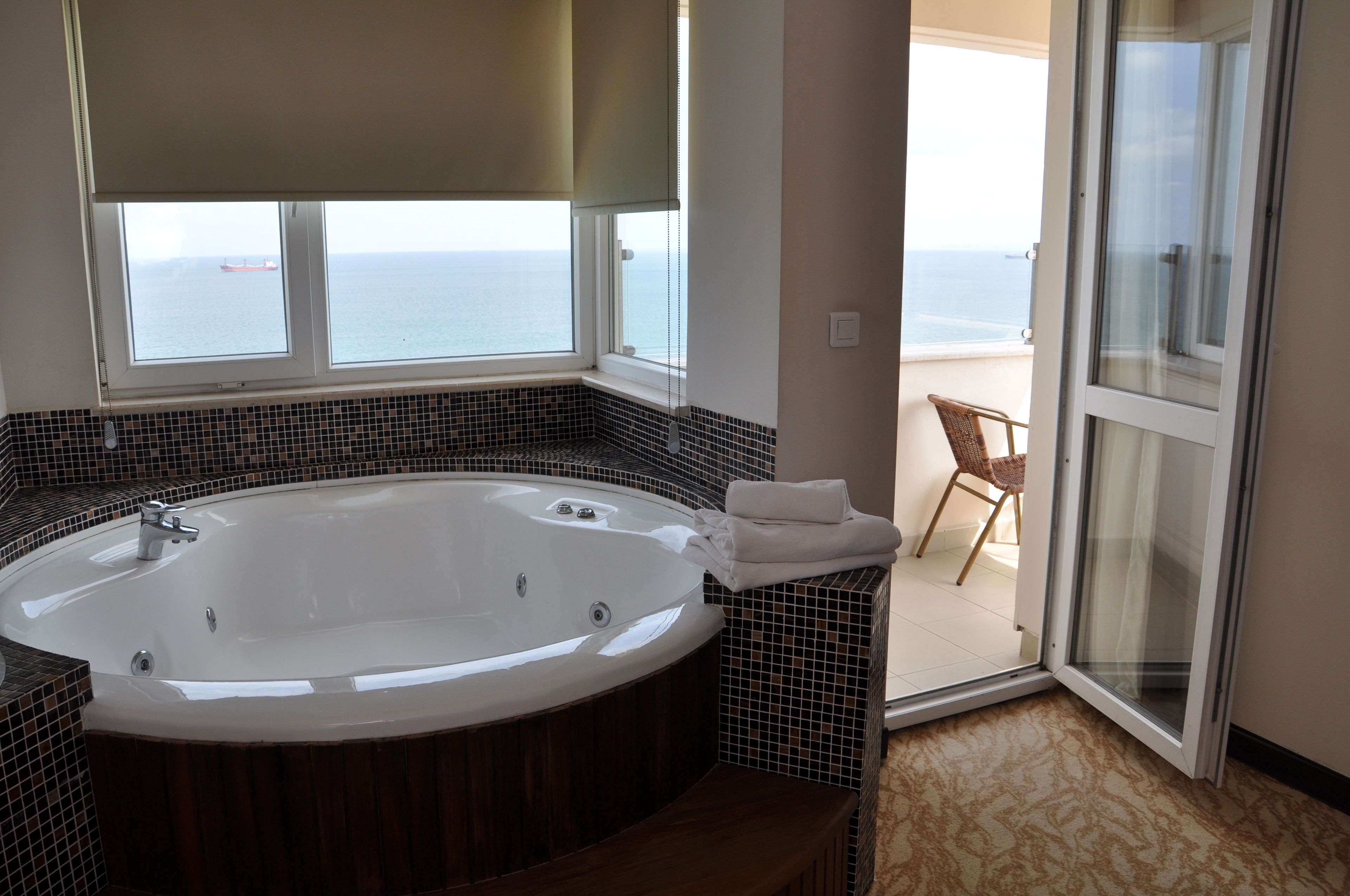 Un baño con una bañera grande, una ventana con vista al mar y una silla en un balcón.