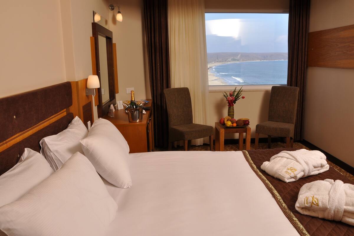 Quarto de hotel com uma cama grande, duas poltronas, uma mesinha com um vaso e frutas, e vista para o oceano.