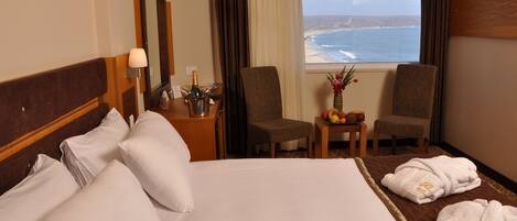Habitación de hotel con una cama grande, dos sillones, una mesita con un florero y fruta, y vista al océano.
