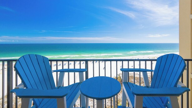 A balcony with two blue Adirondack chairs and a small round table, overlooking a beach with clear blue water and a sandy shore. - Two bedroom Condo 909 at Phoenix 5 (Orange Beach)