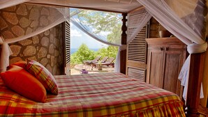 A bed with a plaid bedspread and red pillows, a wooden nightstand, and a view of the outdoors through a window with shutters. - Villa Piton Caribbean Castle | Enhanced cleaning measures. CERTIFIED TO HOST!!! (Soufriere)