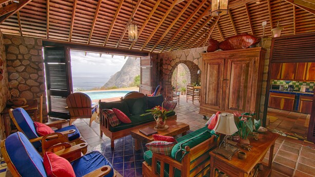 A spacious indoor area with a stone wall, wooden ceiling, and a view of the ocean. There are multiple seating areas with chairs and a sofa. - Villa Piton Caribbean Castle | Enhanced cleaning measures. CERTIFIED TO HOST!!! (Soufriere)