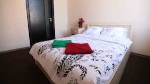 Un lit bien fait, recouvert d’une couvre-lit à motifs floraux, avec une serviette verte et une serviette rouge posées dessus, une petite table de chevet ornée d’un vase rouge contenant une fleur, et une porte fermée.