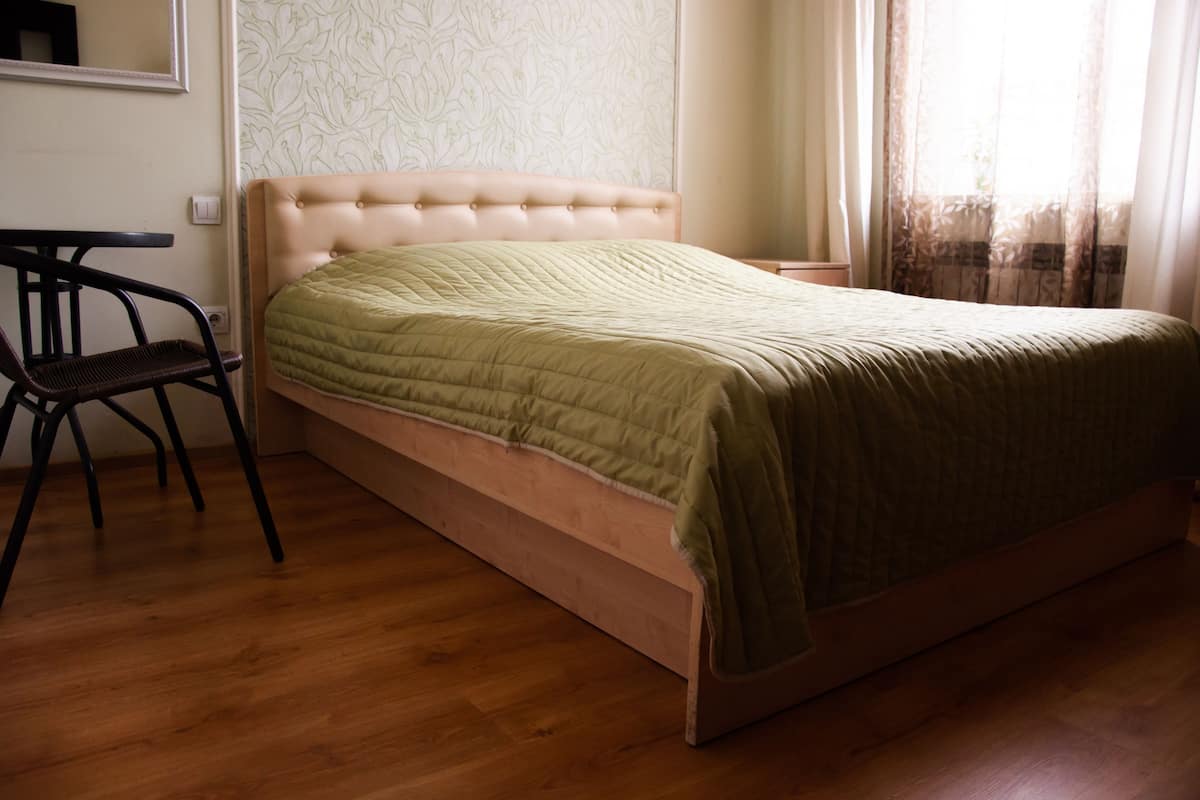 Una cama individual con una colcha verde, una mesita de noche de madera, una silla y un espejo en la pared.
