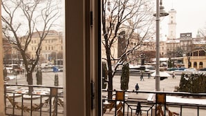 Une vue depuis une fenêtre donnant sur une place de la ville, avec des bâtiments, des arbres et un beffroi.