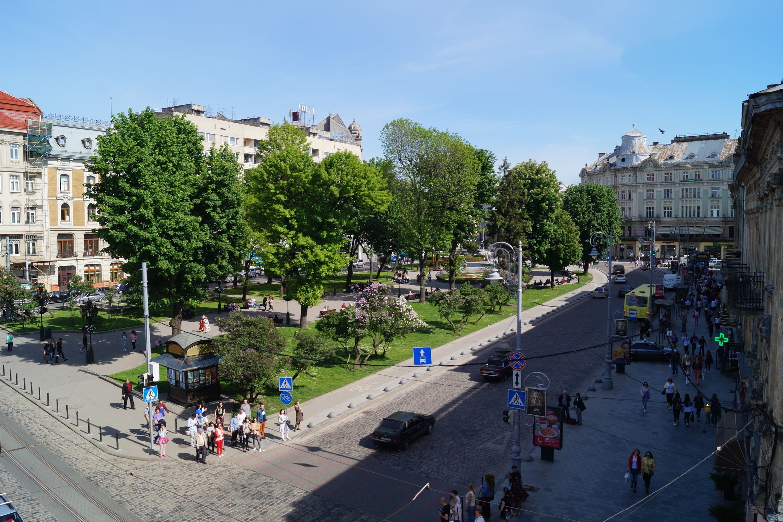 A city square with tram tracks, trees, and people walking.