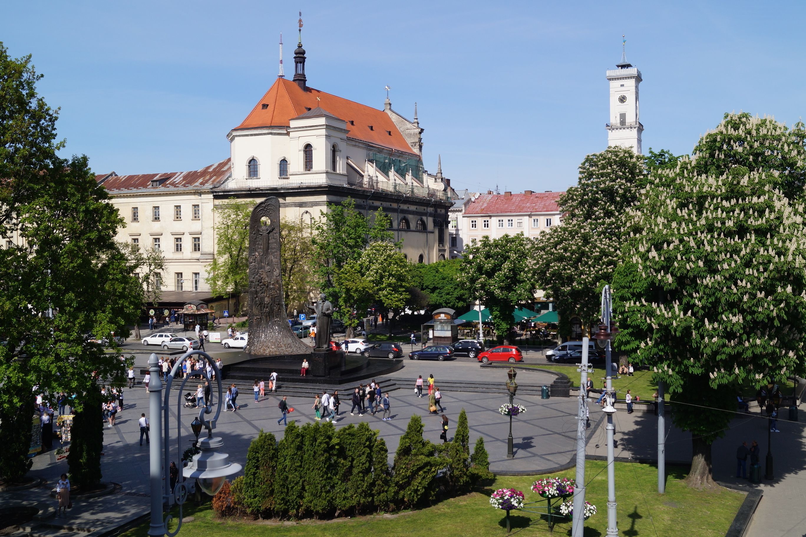 A public square with a prominent building featuring a clock tower, surrounded by trees and a fountain.