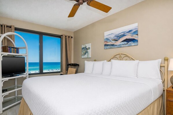 Habitación de hotel con cama, televisor sobre un soporte blanco, ventilador de techo, ventana con vista a la playa y un cuadro en la pared.