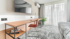 A modern hotel room with a wooden desk, a comfortable chair, a flat-screen TV mounted on the wall, and a large window with sheer curtains. - City Apartments - Apartamenty Młyńska 49 (Koszalin)