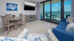 A living room with a blue sofa, a dining table with chairs, a flat-screen TV, and a balcony with a view of the ocean.