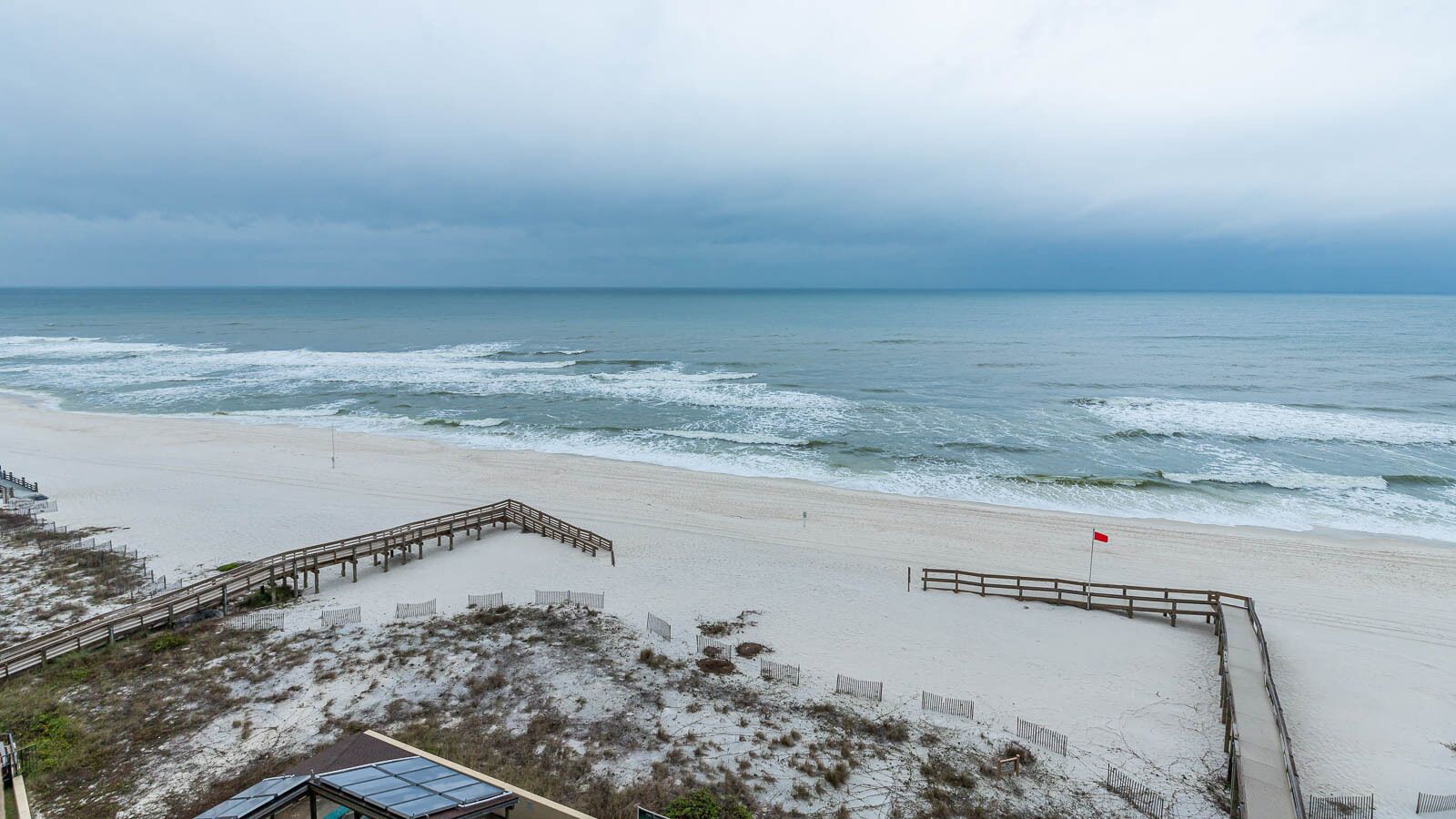 A beach with a wooden walkway leading to a sandy area, a red flag, and a cloudy sky.