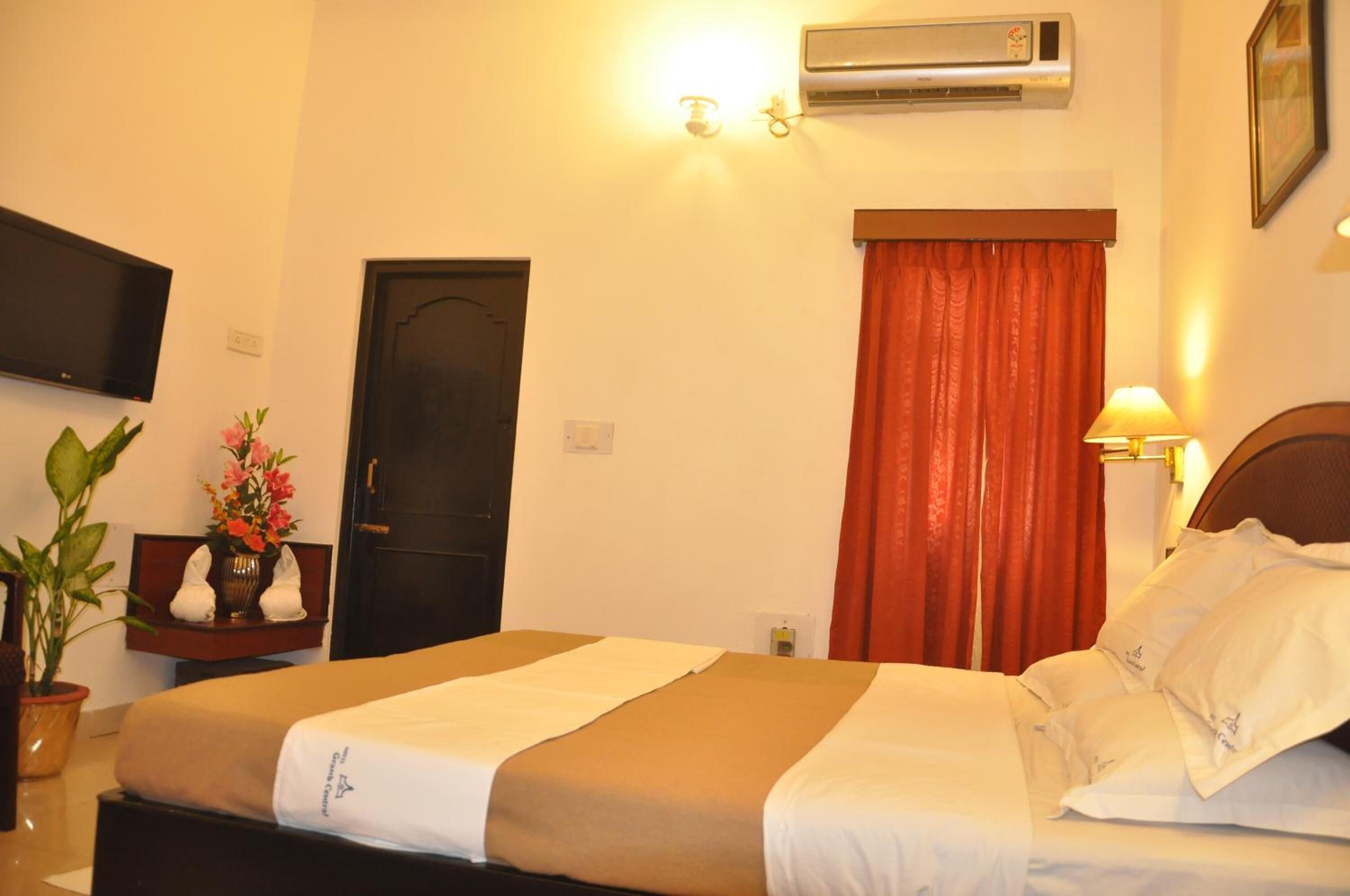 Une chambre d’hôtel avec un lit, un téléviseur fixé au mur, un climatiseur, une porte et une fenêtre avec des rideaux rouges.
