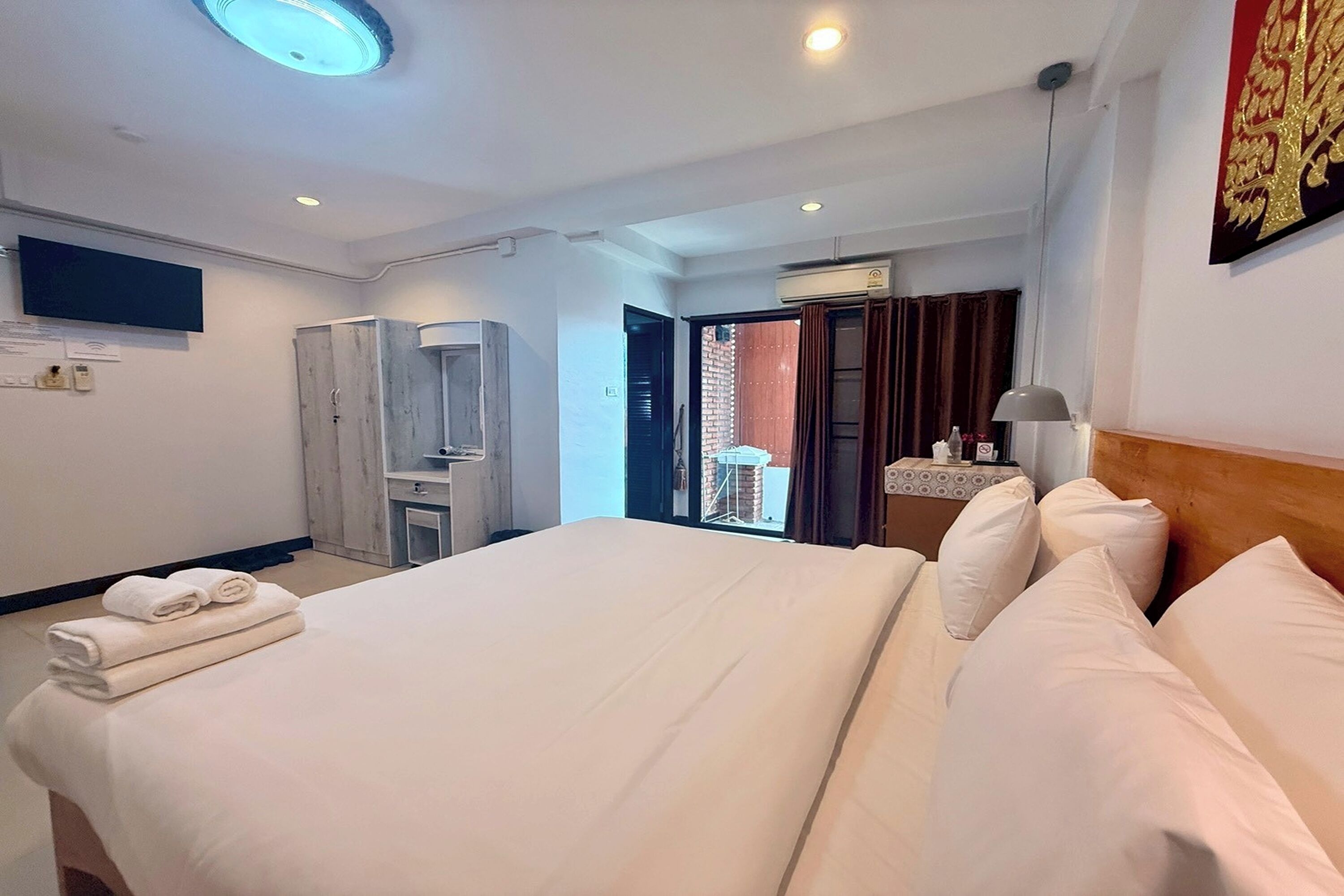 Habitación de hotel con una cama grande, un armario, un escritorio con silla, un televisor montado en la pared y una ventana con cortinas.