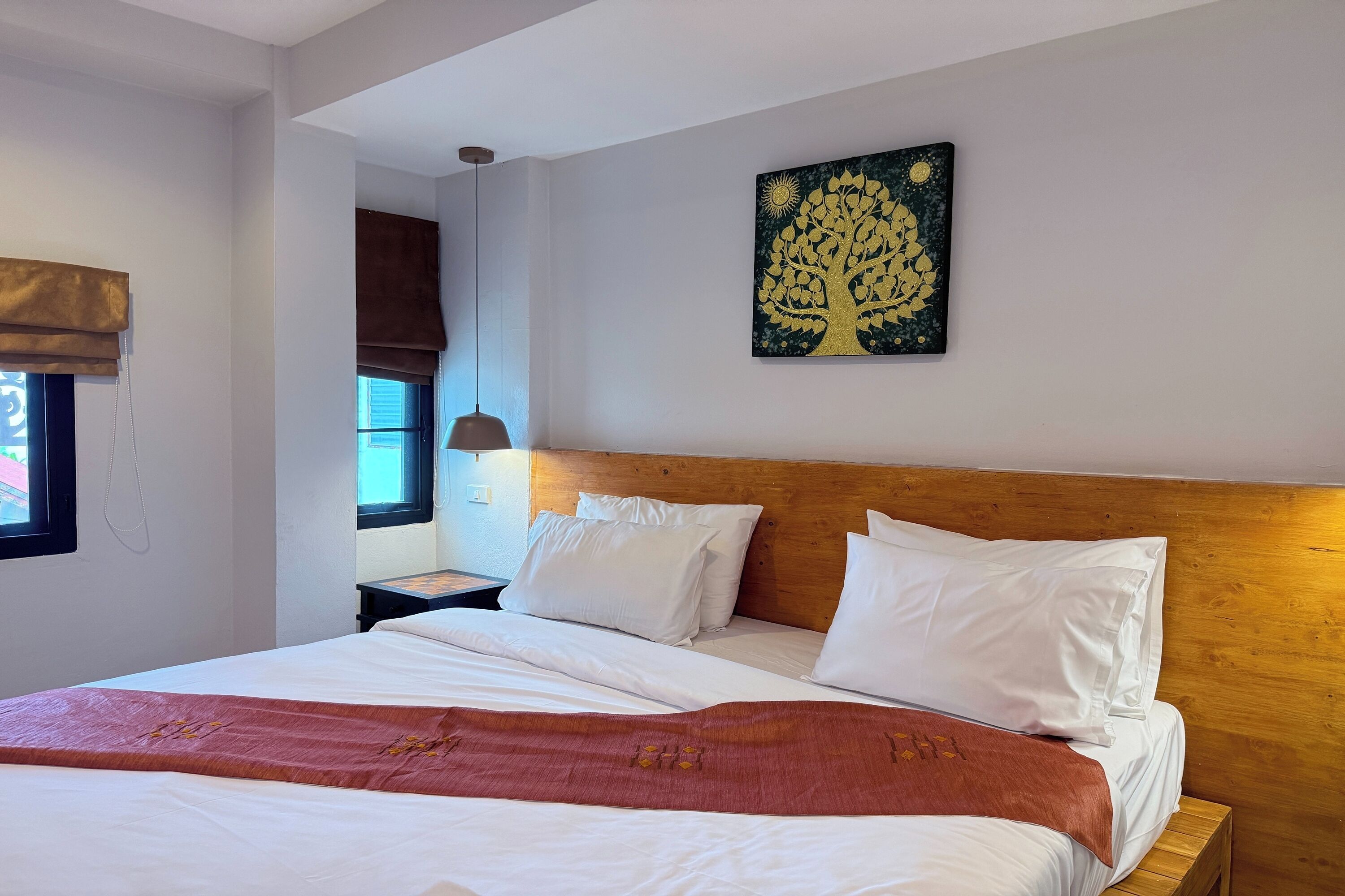 Un lit bien fait, avec du linge de lit blanc et un plaid marron, une tête de lit en bois et une œuvre d’art représentant un arbre accrochée au mur.
