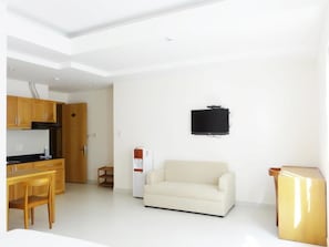 Una sala de estar moderna con un sofá, una mesa de centro, un televisor montado en la pared y una zona de cocina con armarios y un refrigerador.