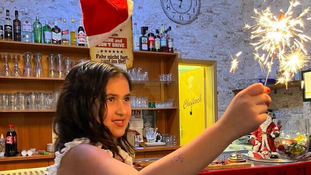 Eine Person hält ein Feuerwerk in einer Bar mit einer Ziegelwand, Regalen voller Flaschen und einer Uhr.