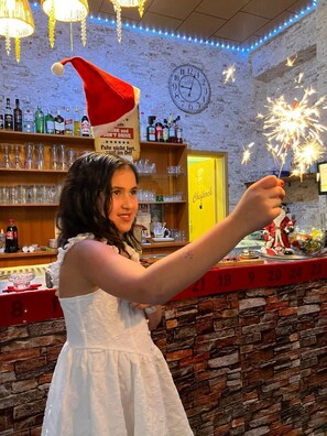 Eine Person hält ein Feuerwerk in einer Bar mit einer Ziegelwand, Regalen voller Flaschen und einer Uhr.