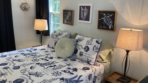 Une chambre à coucher avec un lit agrémenté de linge de lit sur le thème de l’océan, une table de chevet avec une lampe et, au mur, des œuvres d’art encadrées représentant des hippocampes.