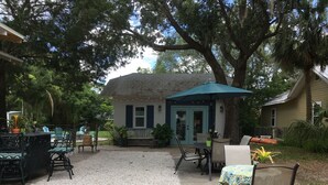 Un espace extérieur aménagé en terrasse avec des tables et des chaises, un coin salon abrité et un bâtiment doté d’une porte et de fenêtres bleues.