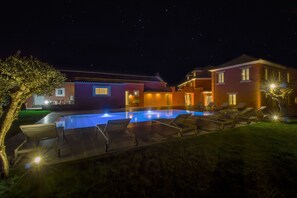 A nighttime view of a hotel with a swimming pool, lounge chairs, and well-lit buildings.