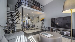 A modern living room with a grey sofa, a round ottoman, a large flat-screen TV, and a staircase leading to a loft. - Paço do Bispo Boutique House (Cascais)