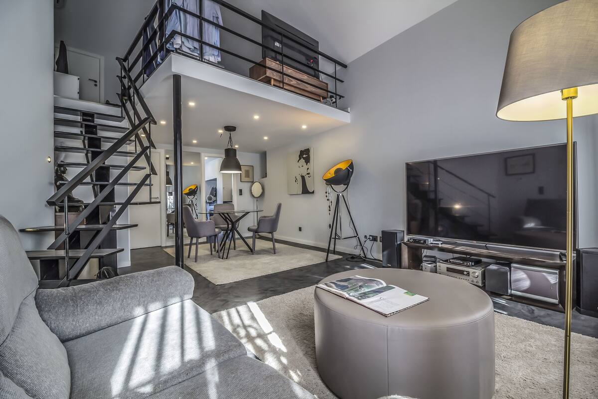 Een moderne woonkamer met een grijze bank, een rond hockertje, een grote flatscreen-televisie en een trap naar een vide.
