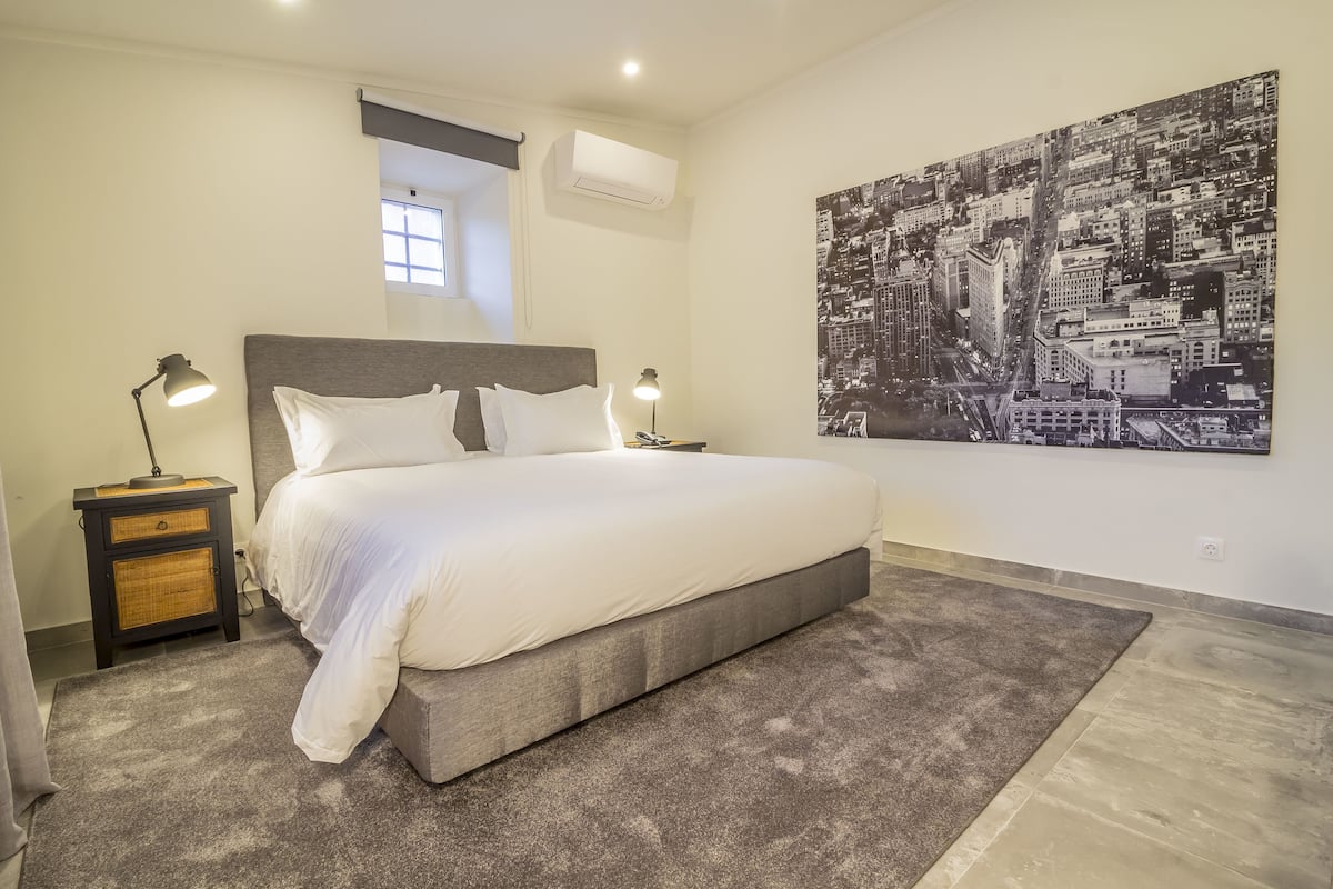 Een slaapkamer met een groot bed, twee nachtkastjes met lampen, een raam, een airconditioner en een stadslandschap schilderij aan de muur.