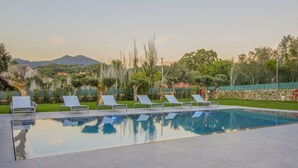 A rectangular swimming pool with clear water, surrounded by a tiled deck. There are several white lounge chairs aligned along the pool's edge. The pool is set against a backdrop of trees and a distant mountain range. - Paço do Bispo Boutique House (Cascais)