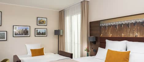 A hotel room with two beds, a large window with curtains, a wall-mounted TV, and framed pictures on the wall.