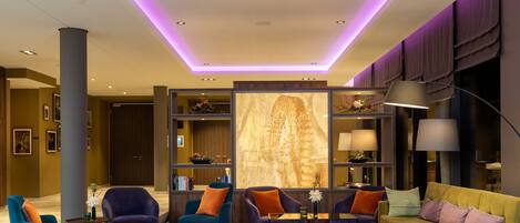 Un vestíbulo moderno con techo alto, iluminación empotrada y una pared decorativa central. Hay zonas de estar con sillones y un sofá, así como una estantería con diversos objetos.