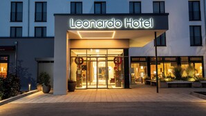 El exterior del Hotel Leonardo, con una fachada moderna, una entrada de vidrio amplia y un área de acceso bien iluminada.