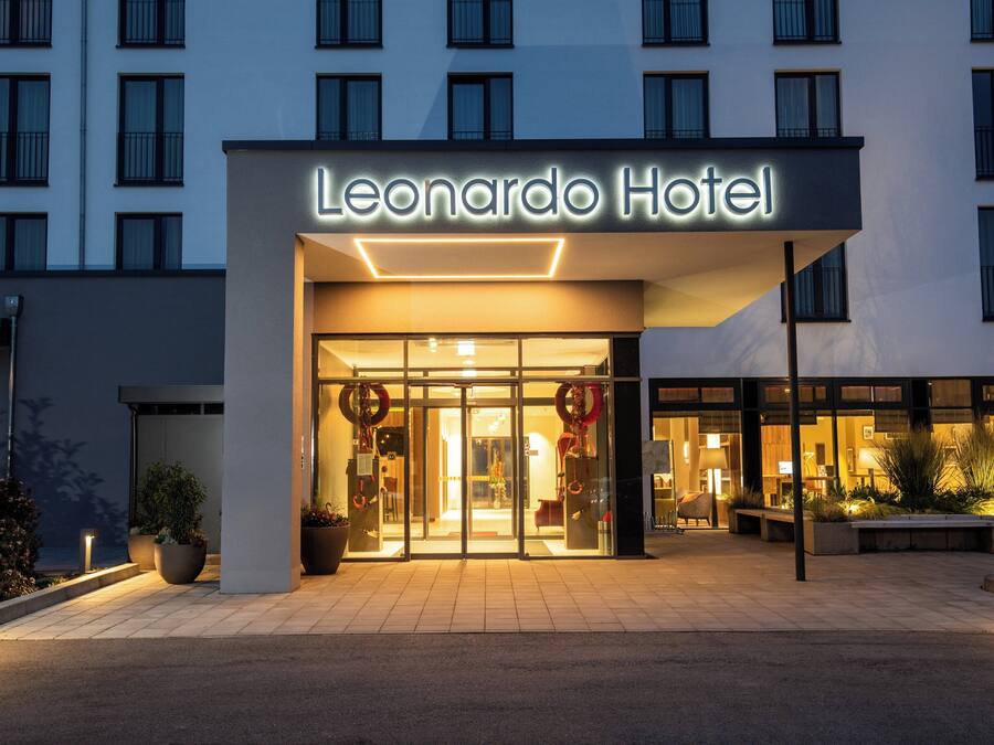El exterior del Hotel Leonardo, con una fachada moderna, una entrada de vidrio amplia y un área de acceso bien iluminada.
