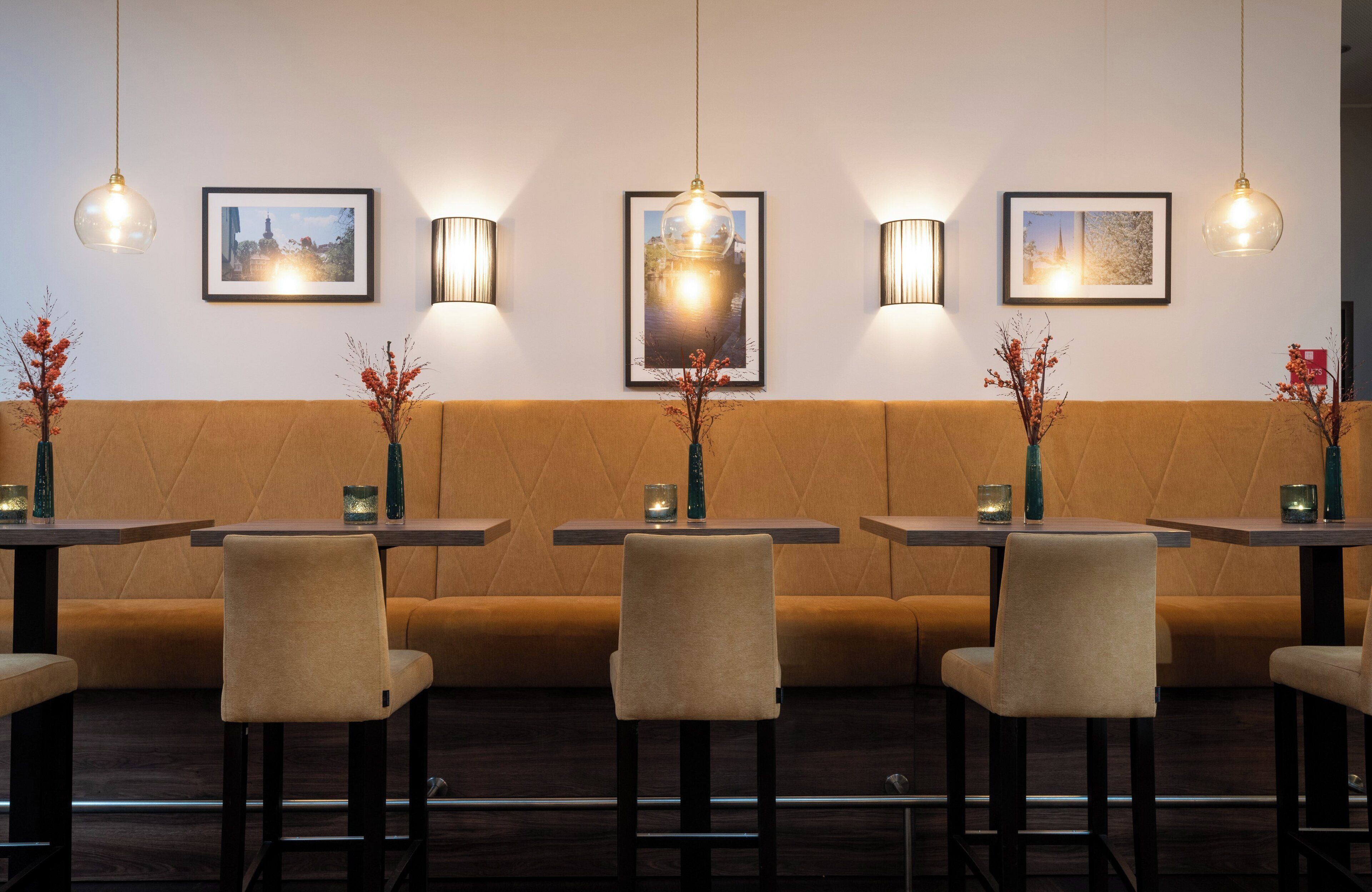 Interior de restaurante con un largo banco acolchado marrón, varias mesas con sillas, lámparas colgantes e imágenes enmarcadas en la pared.