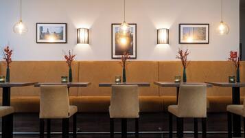 Interior de restaurante con un largo banco acolchado marrón, varias mesas con sillas, lámparas colgantes e imágenes enmarcadas en la pared.