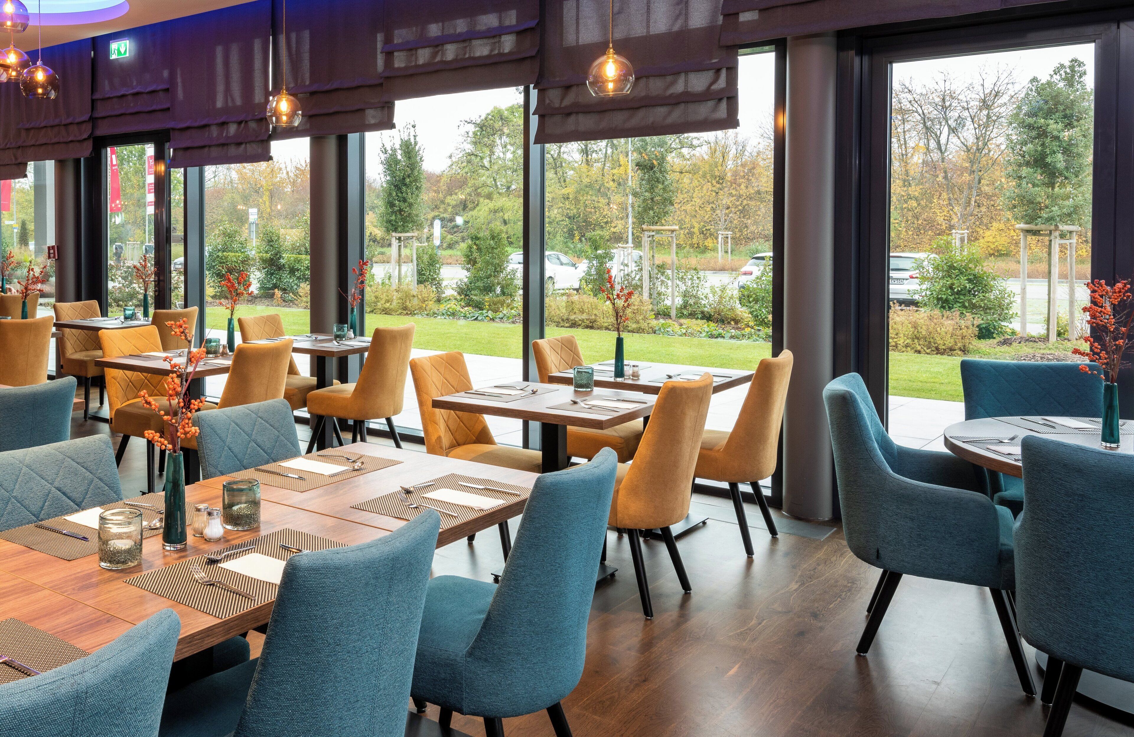 Un restaurant au décor moderne, avec des tables en bois et des chaises bleues, de grandes fenêtres offrant une vue sur l’extérieur.