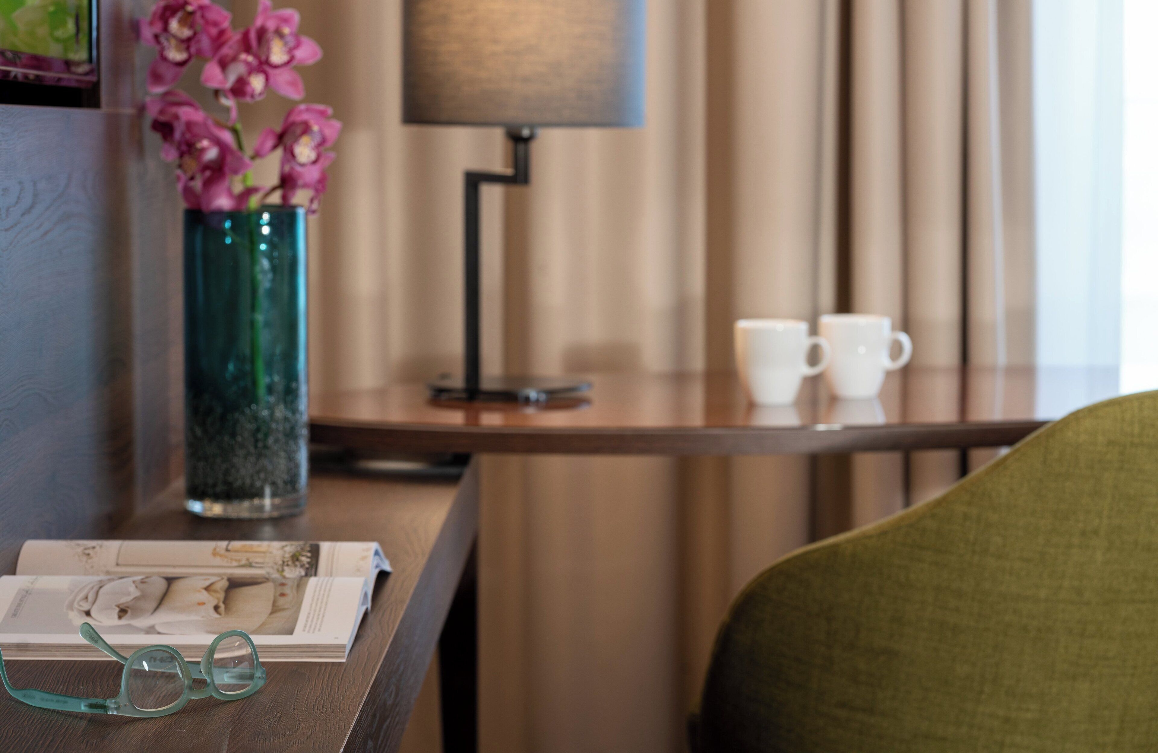 A wooden table with a vase of pink flowers, a lamp, two white cups, and a pair of glasses on a magazine.